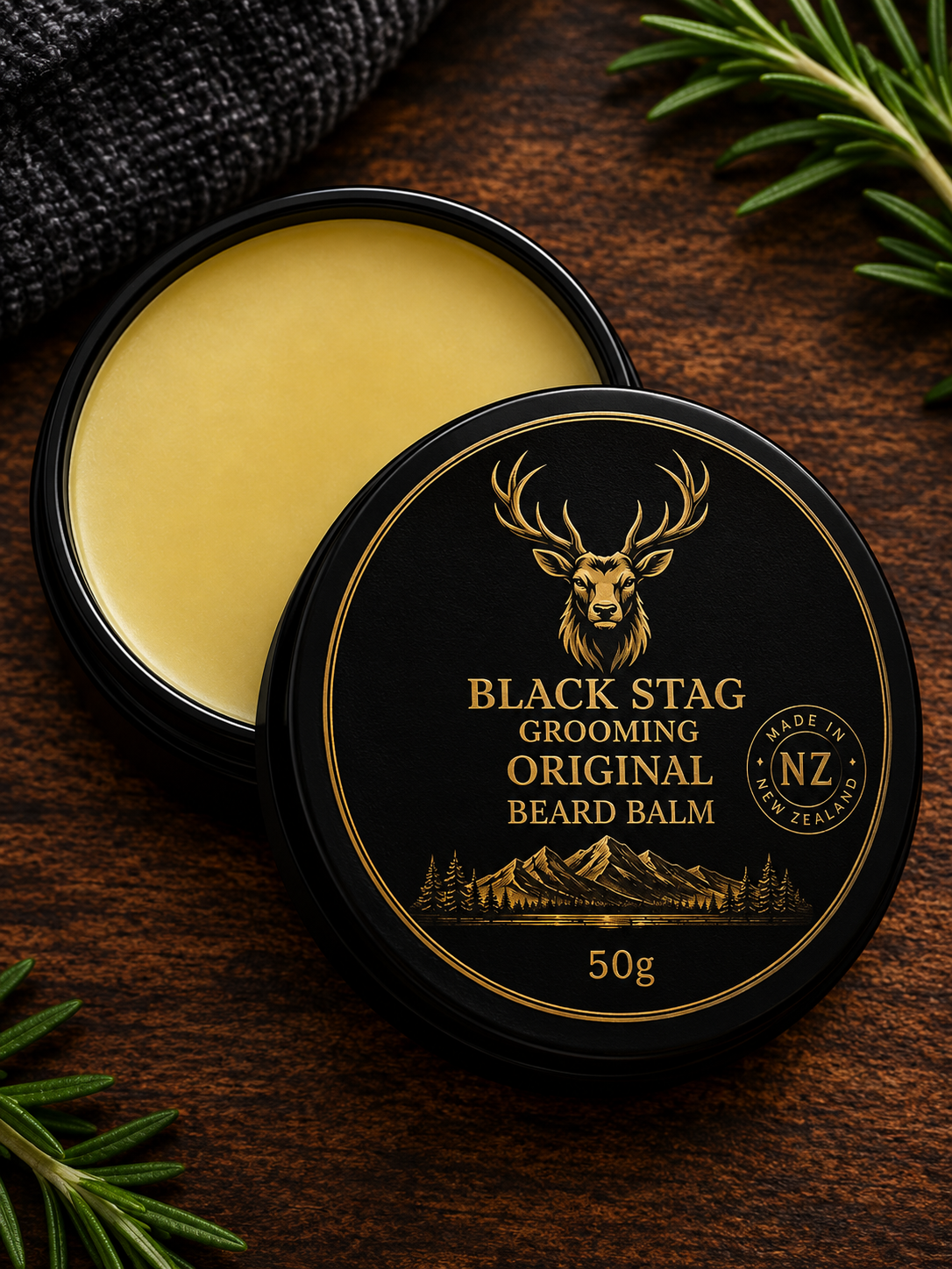 An open round black tin of Black Stag Grooming Original Beard Balm, showing the pale yellow balm inside, with the lid placed in front. The lid has a gold stag head logo and mountain illustration.