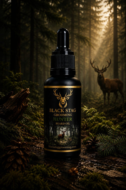 A bottle of Black Stag Grooming Hunter Beard Oil stands in a dark forest setting with a stag in the background. The bottle is black with gold lettering and a deer head logo, set against a moody, misty woodland scene.