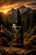 A bottle of Black Stag Grooming Original Beard Oil stands on a rustic log outdoors, with mountains, pine trees, and a glowing sunset in the background. The bottle is black with gold lettering and a deer logo, surrounded by pinecones and moss.