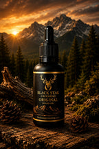 A bottle of Black Stag Grooming Original Beard Oil stands on a rustic log outdoors, with mountains, pine trees, and a glowing sunset in the background. The bottle is black with gold lettering and a deer logo, surrounded by pinecones and moss.